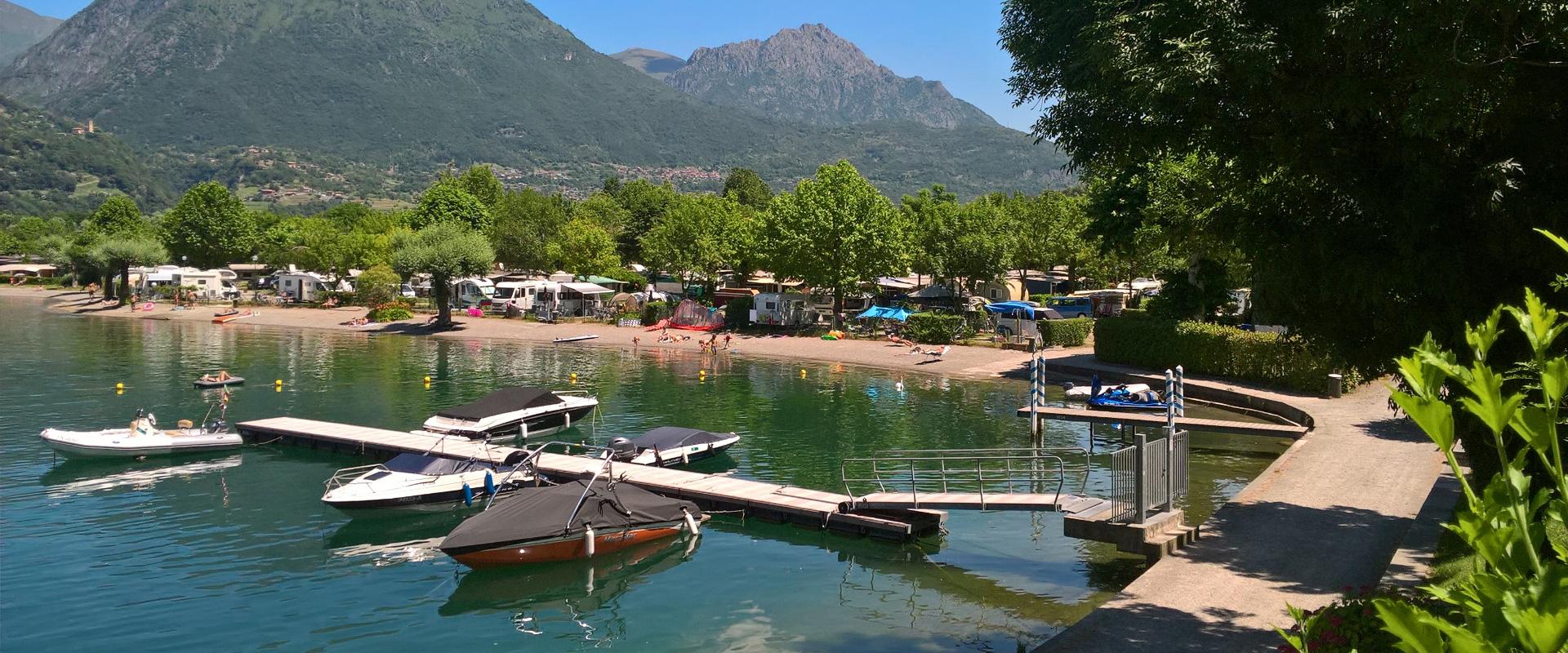 Campeggio sul lago con barche, montagne e natura rigogliosa.