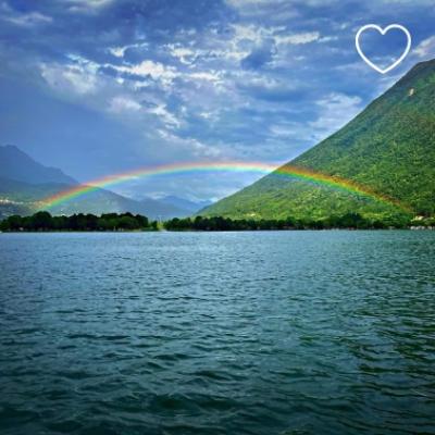 Arcobaleno sopra un lago circondato da montagne verdi e cielo nuvoloso.