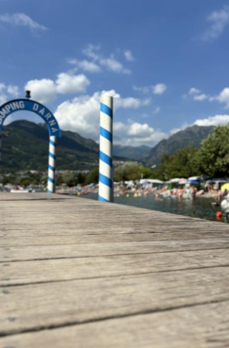 Spiaggia lacustre con barche, montagne verdi e campeggio, giornata estiva soleggiata.