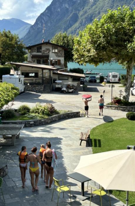 Campeggio sul lago con persone che si dirigono verso l'acqua, montagne sullo sfondo.