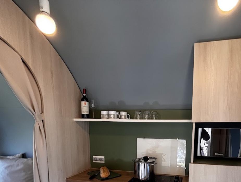 Piccola cucina con fornello, microonde, frigo, caffettiera e mensole con bicchieri.