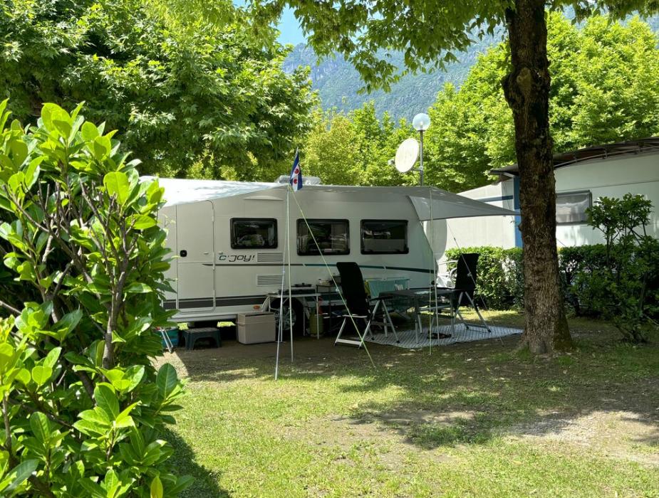 Roulotte parcheggiata in campeggio, circondata da alberi e sedie da giardino.