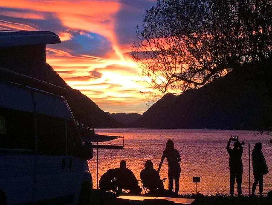 Tramonto colorato su lago, persone e camper in silhouette.