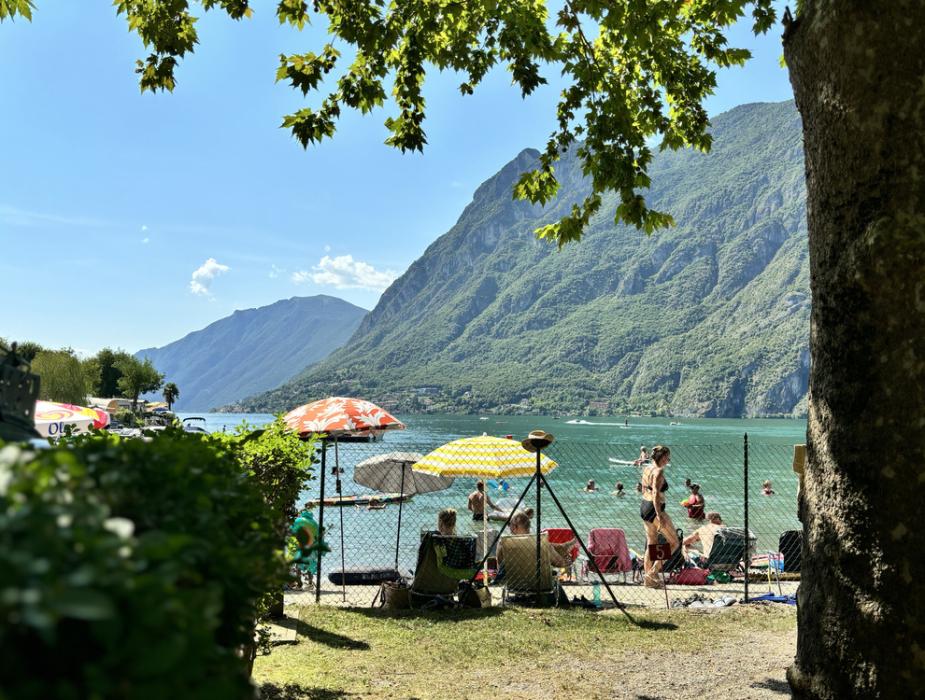 Spiaggia affollata sul lago, ombrelloni colorati e montagne verdi sullo sfondo.