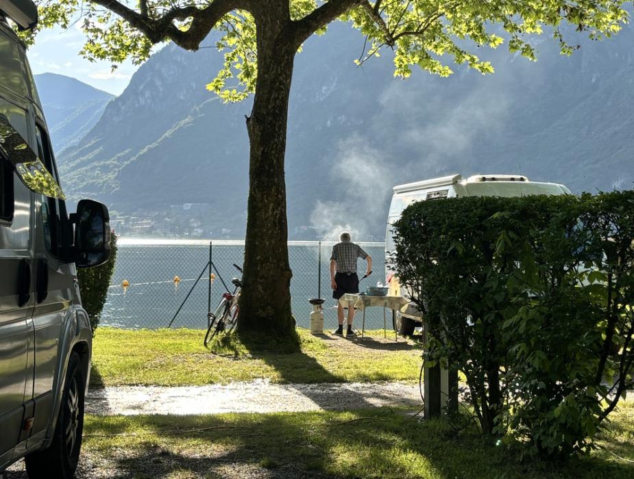Campeggio sul lago, uomo cucina all'aperto sotto un albero, vista monti.