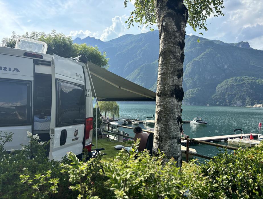 Camper parcheggiato vicino al lago con vista sulle montagne e barche.