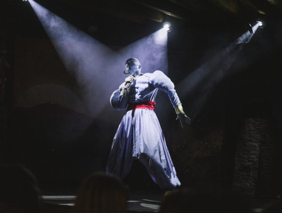 Attore sul palco in costume da genio, illuminato da luci teatrali.