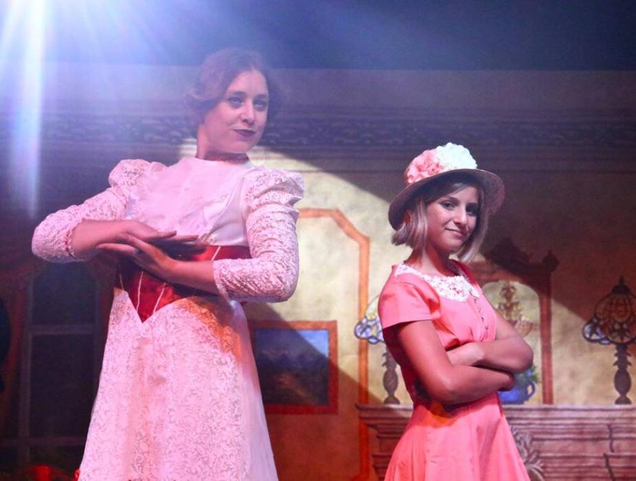 Due attrici in scena con costumi d'epoca, illuminate da una luce teatrale.