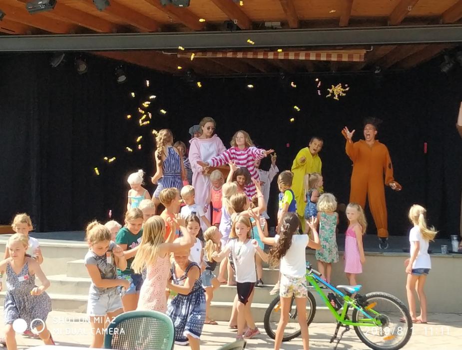 Bambini sul palco con animatori in costume, lanciando caramelle, durante un evento all'aperto.