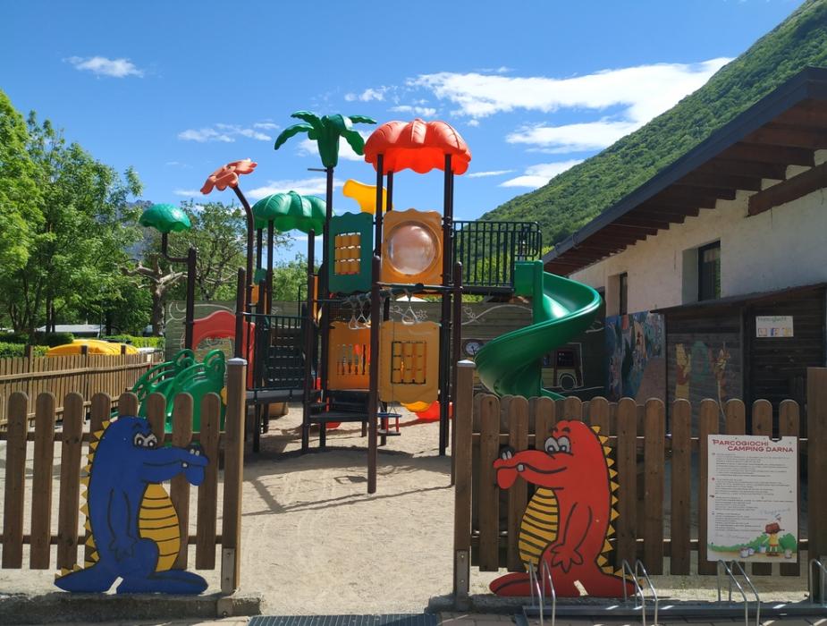 Parco giochi colorato con scivoli e decorazioni di coccodrilli, circondato da montagne verdi.